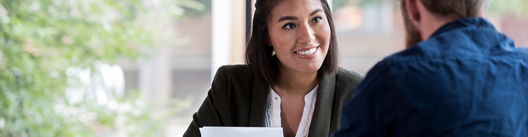 Image of a young woman smiling at another person while holding a piece of white paper.