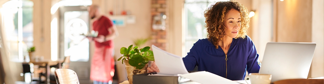 Image of a woman in a cafe looking over paperwork while working on her laptop.